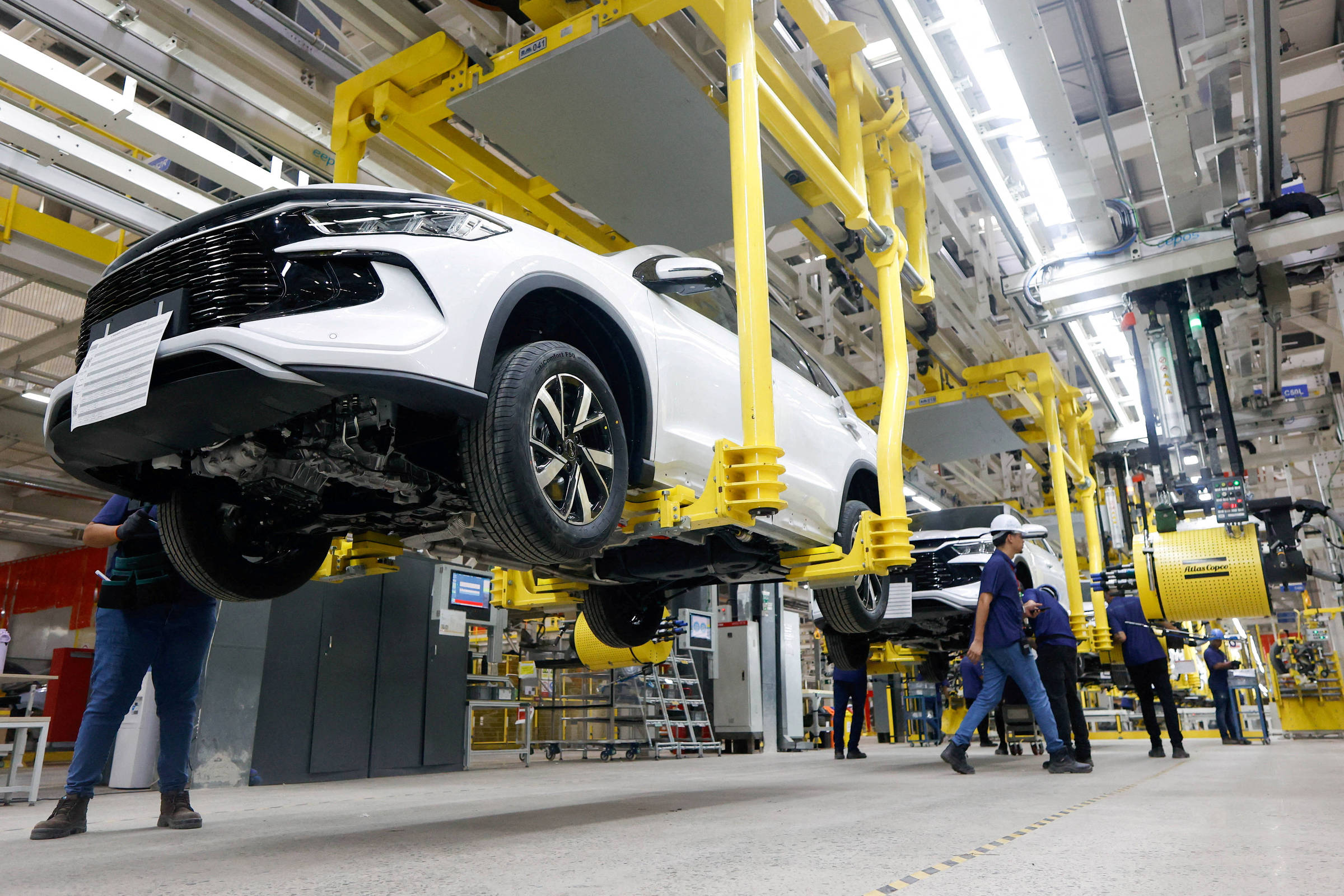 Carro branco suspenso por estrutura amarela em linha de montagem dentro de fábrica. Trabalhadores com uniformes e capacetes circulam pelo ambiente industrial com equipamentos e maquinário ao fundo.