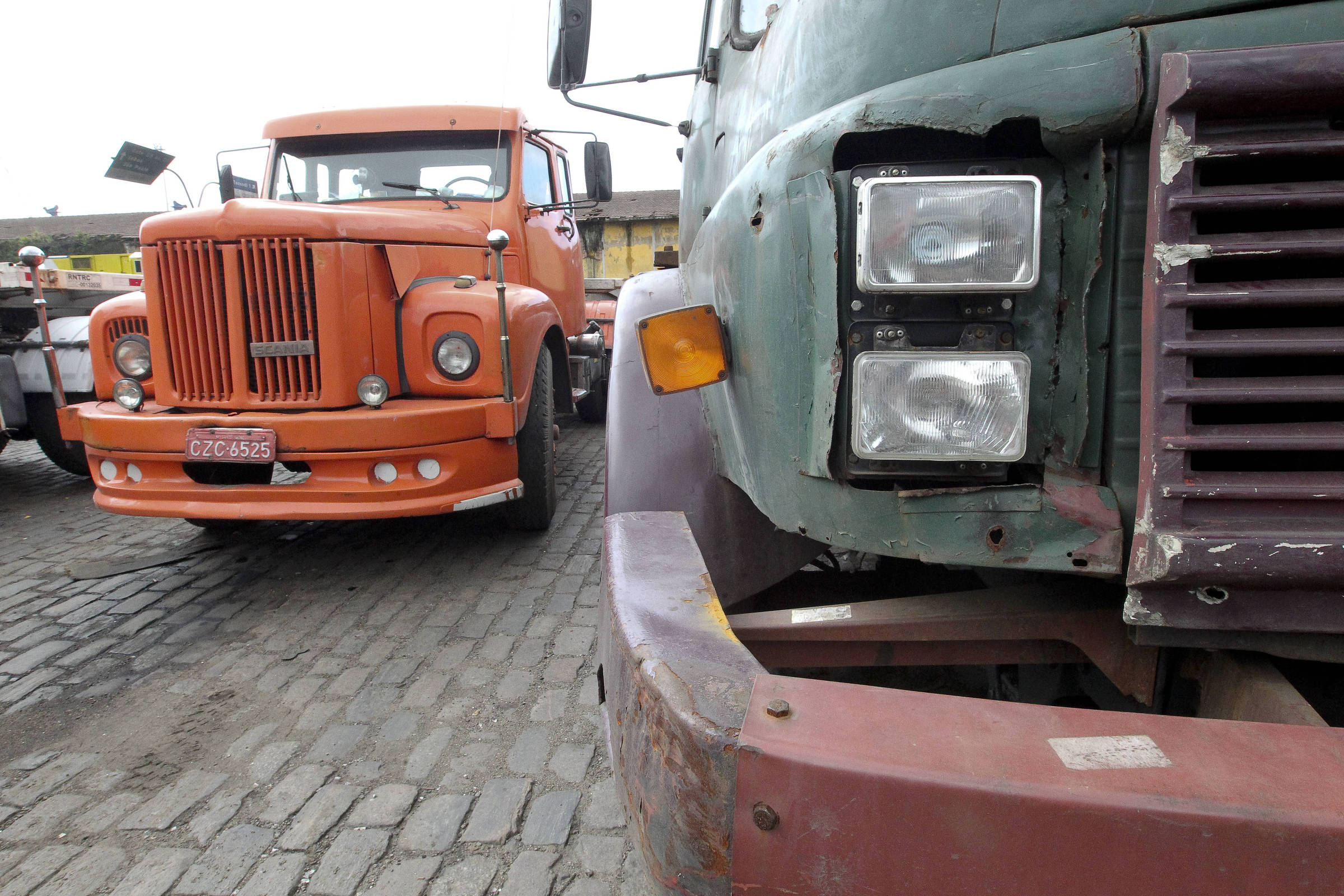 Dois caminhões antigos estacionados lado a lado em pátio com piso de paralelepípedos. O caminhão à esquerda é laranja, com grade frontal vertical e placa CZC-6525. O caminhão à direita está parcialmente visível, com pintura verde desgastada, faróis quadrados duplos e para-choque enferrujado.